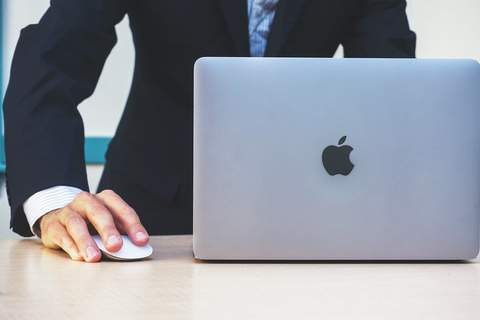 A hand resting on a wireless mouse next to a laptop on a desk, representing the day-to-day work of researching and writing web content